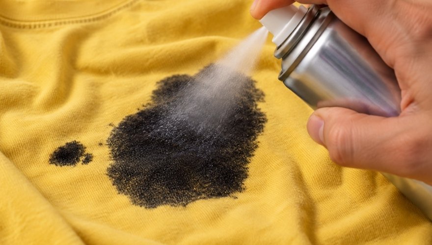 Hairspray is sprayed onto a large black ink stain on a yellow shirt.