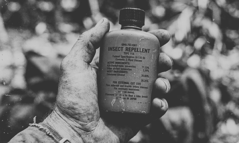 A weathered hand holds a small bottle labeled “Insect Repellent” against blurred foliage