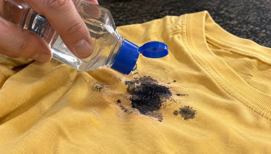 Clear liquid is poured onto a black ink stain on a yellow shirt.
